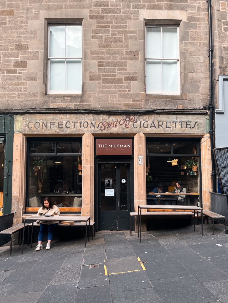 a stone building on a cobbled street in Edinburgh. There is a coffee shop in the bottom unit that looks slightly aged with time and has old signage that has faded. A couple sit at the window and another woman sits outside on a bench.