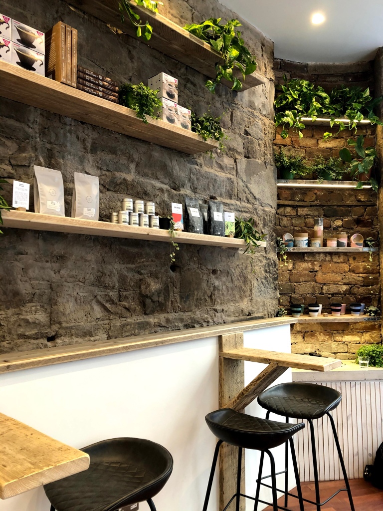 The interior of a coffee shop in Edinburgh with exposed brick walls. There is an industrial aesthetic with wooden planks being used as bar tables with modern leather stools tucked underneath. Similar planks have also been used as shelving for reusable coffee cups and plants. 